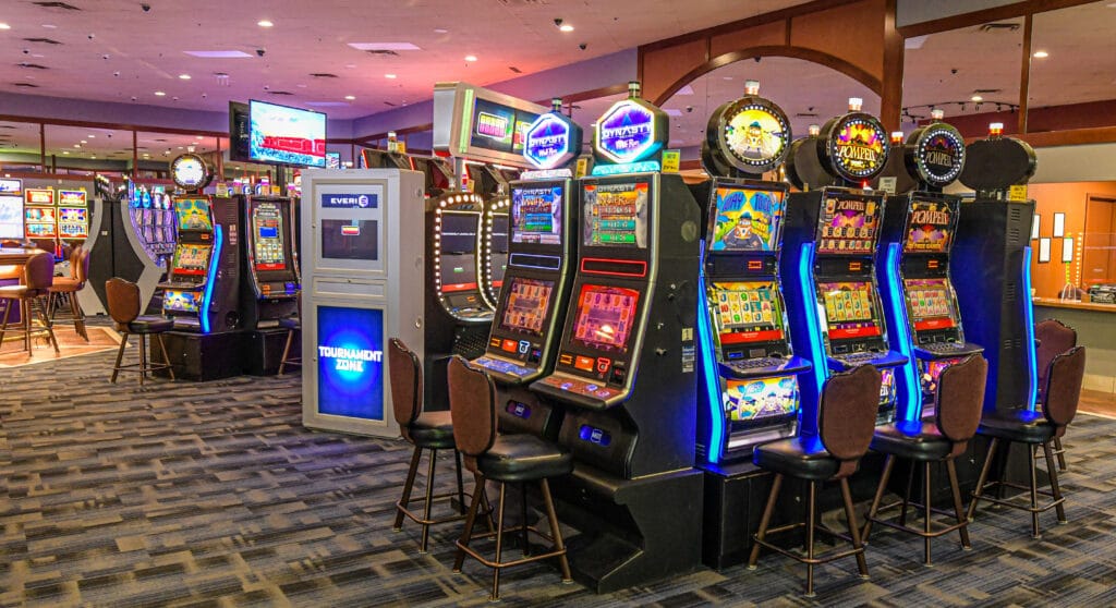 Numerous brightly lit slot machines with stools are arranged in rows on a patterned carpet inside a casino.