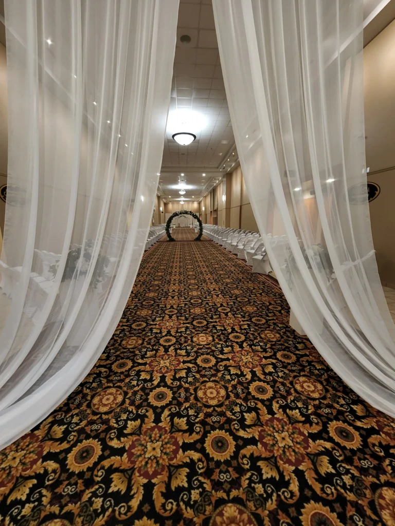 Sheer white curtains frame a long, patterned carpeted aisle, flanked by rows of white chairs, that leads toward a decorated archway.
