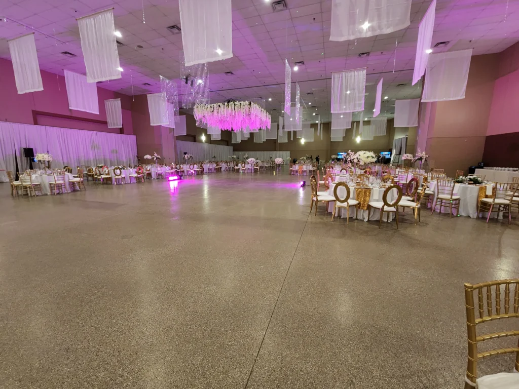 A large banquet hall is elegantly set with numerous round tables and gold chairs, featuring white drapes along the walls, hanging fabric panels from the ceiling, and a central floral chandelier illuminated by purple light.