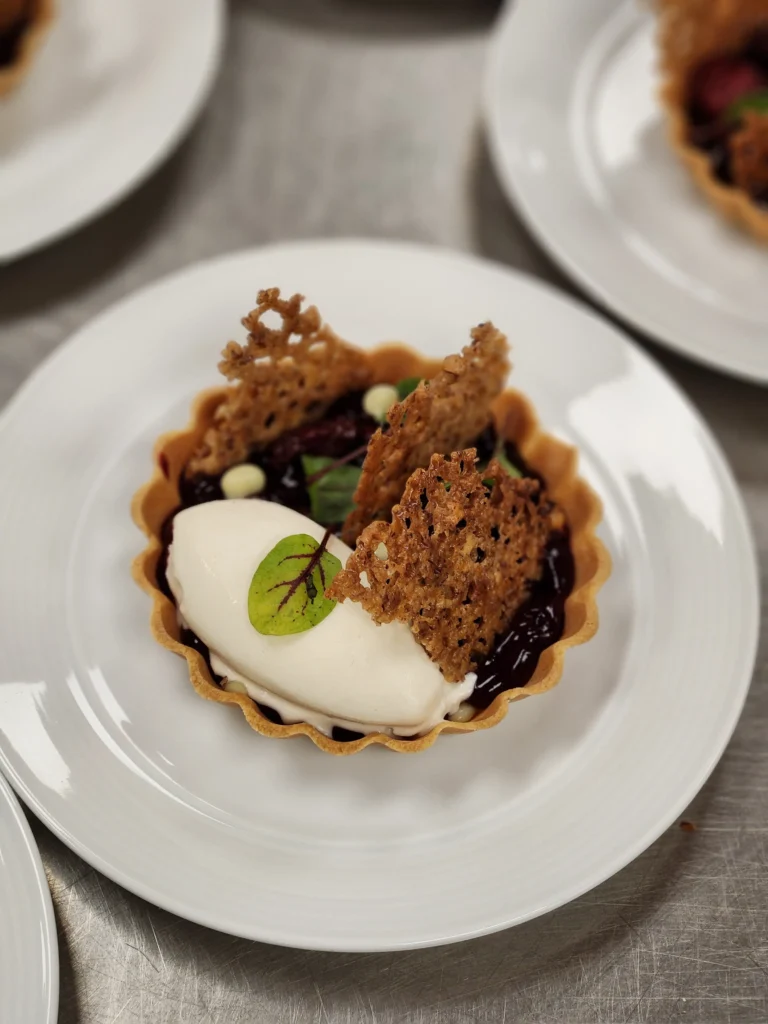 A dessert tart with a dark berry filling, a scoop of white ice cream garnished with a small green leaf, and crispy wafers is presented on a white plate, with other desserts blurred in the background on a metal counter.