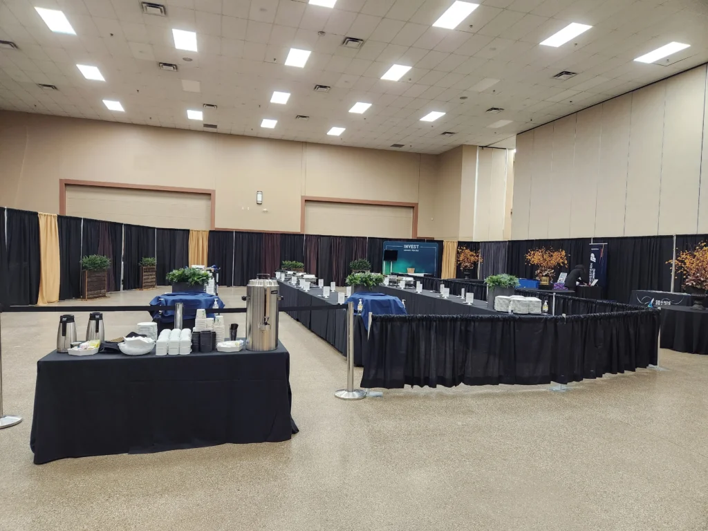 An expansive room is prepared for an event with long black draped tables for beverages and food service, backed by black and golden fabric panels, all beneath a bright white drop ceiling.