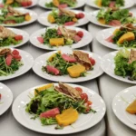 Rows of white plates display colorful salads made with mixed greens, red grapefruit segments, golden beet slices, roasted cherry tomatoes, and folded flatbreads on a light gray table.
