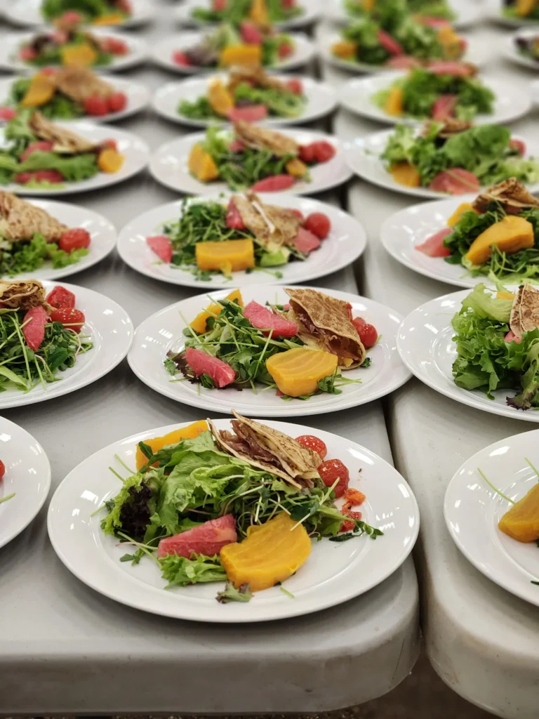 Many white plates are neatly arranged across a surface, each holding a colorful salad made of mixed greens, golden beets, pink grapefruit, cherry tomatoes, and a folded crepe.