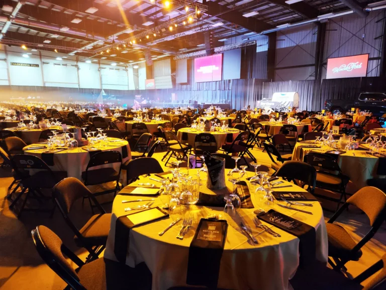 The vast event hall is meticulously prepared for a large gathering, featuring countless round tables adorned with full place settings, illuminated by stage lights, and backed by large display screens, a covered wagon, and a truck.