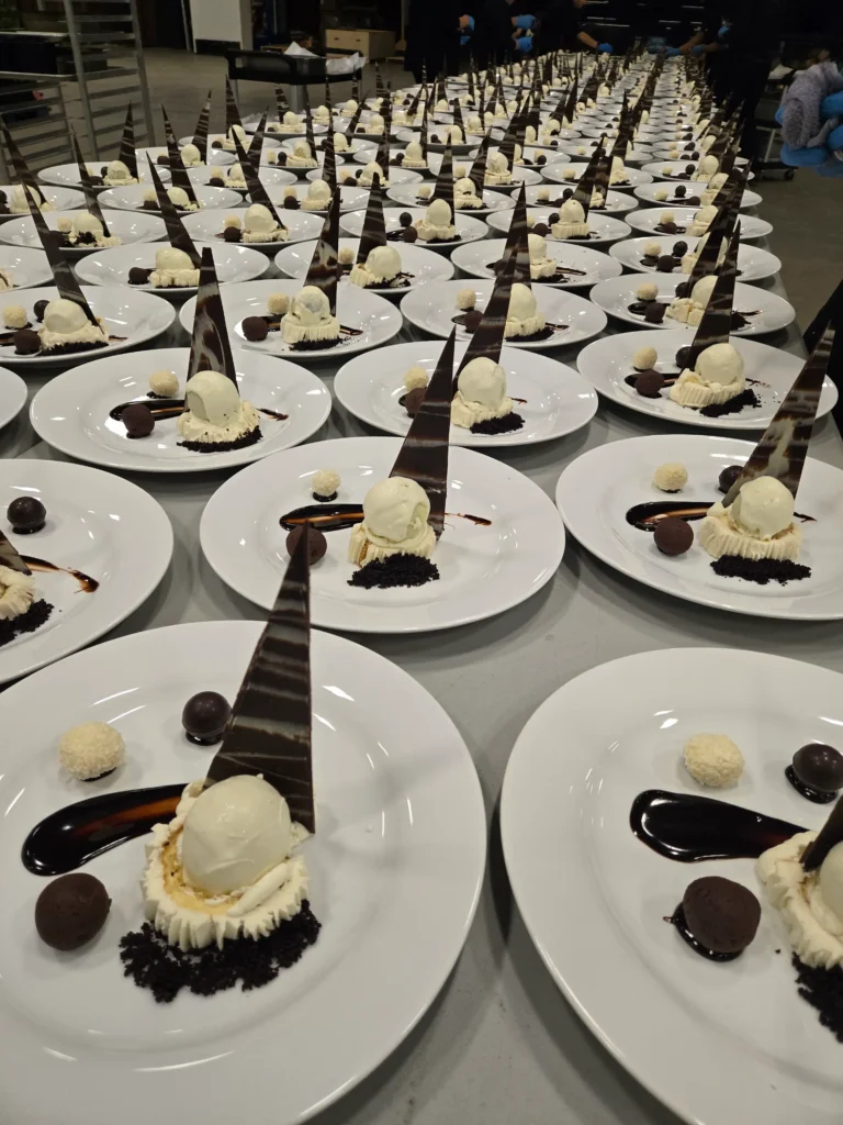 Rows of identical dessert plates, each featuring a scoop of white dessert atop dark crumbs, a tall patterned chocolate triangle, chocolate syrup, and two small round candies, are neatly displayed on a table while people work in the blurred background.