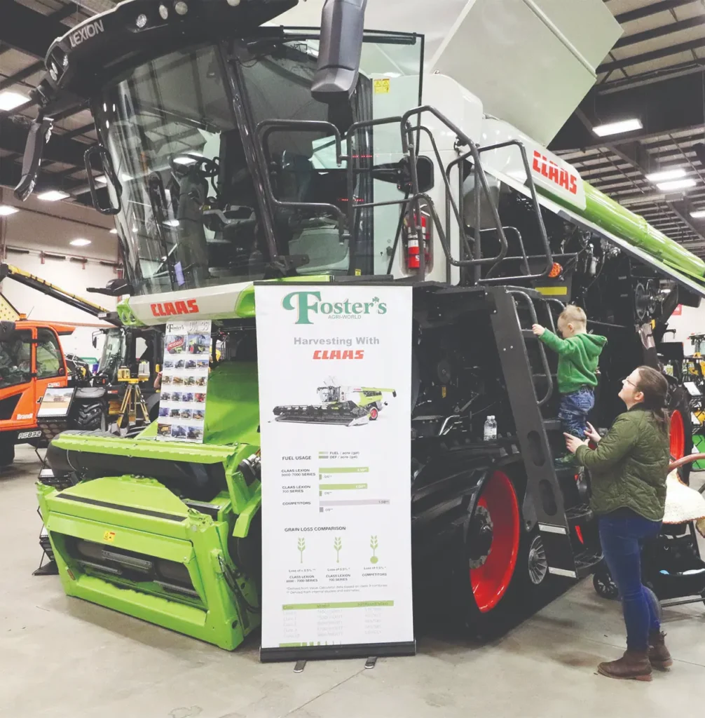 A child climbs a large green Claas combine harvester as a woman watches nearby, positioned next to a Fosters Agri World promotional banner.