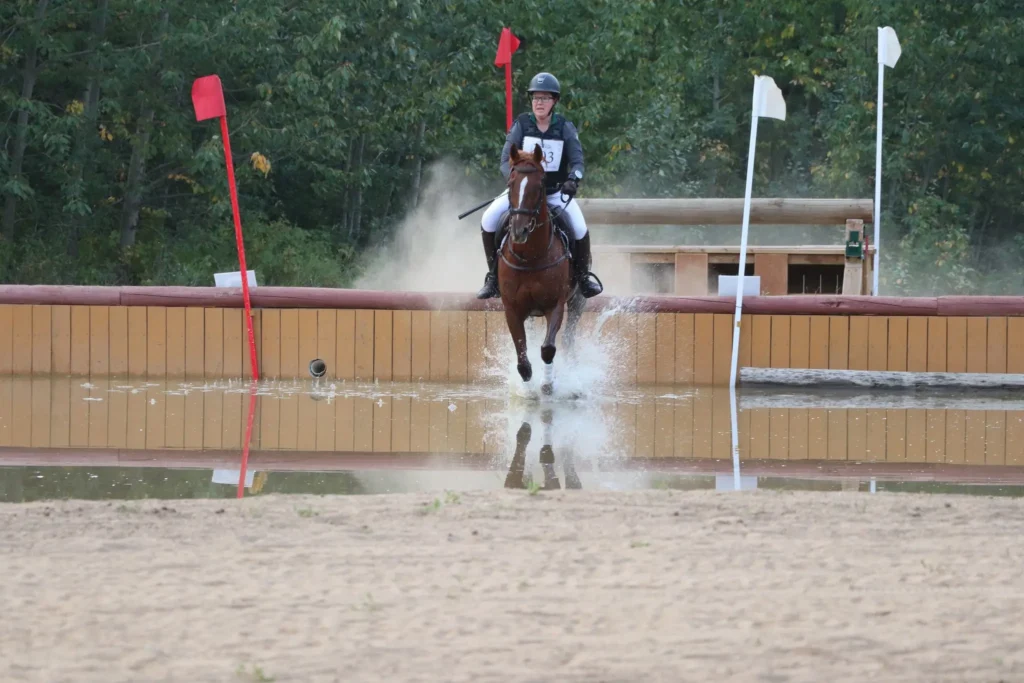 A rider wearing a helmet and a number thirteen bib guides a brown horse through a water obstacle, creating a large splash between red and white course flags, with green trees in the background.