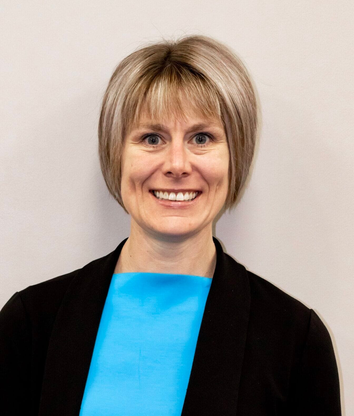 A smiling woman with short blonde hair and blue eyes wears a black blazer over a bright blue top against a plain light background.