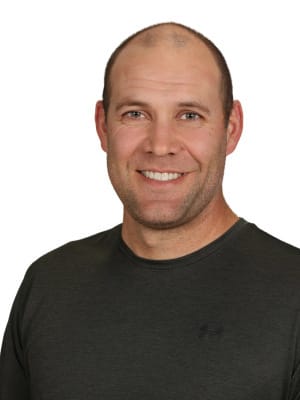 A man with a short haircut and a dark shirt smiles towards the viewer against a plain white background.