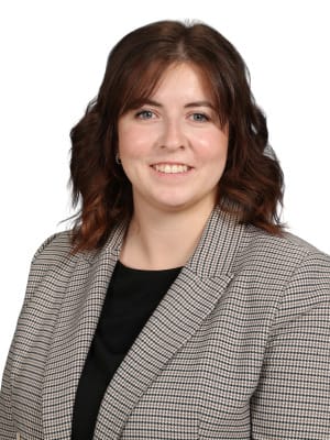 A smiling woman with wavy brown hair and bangs wears a plaid blazer over a black top against a plain white background.