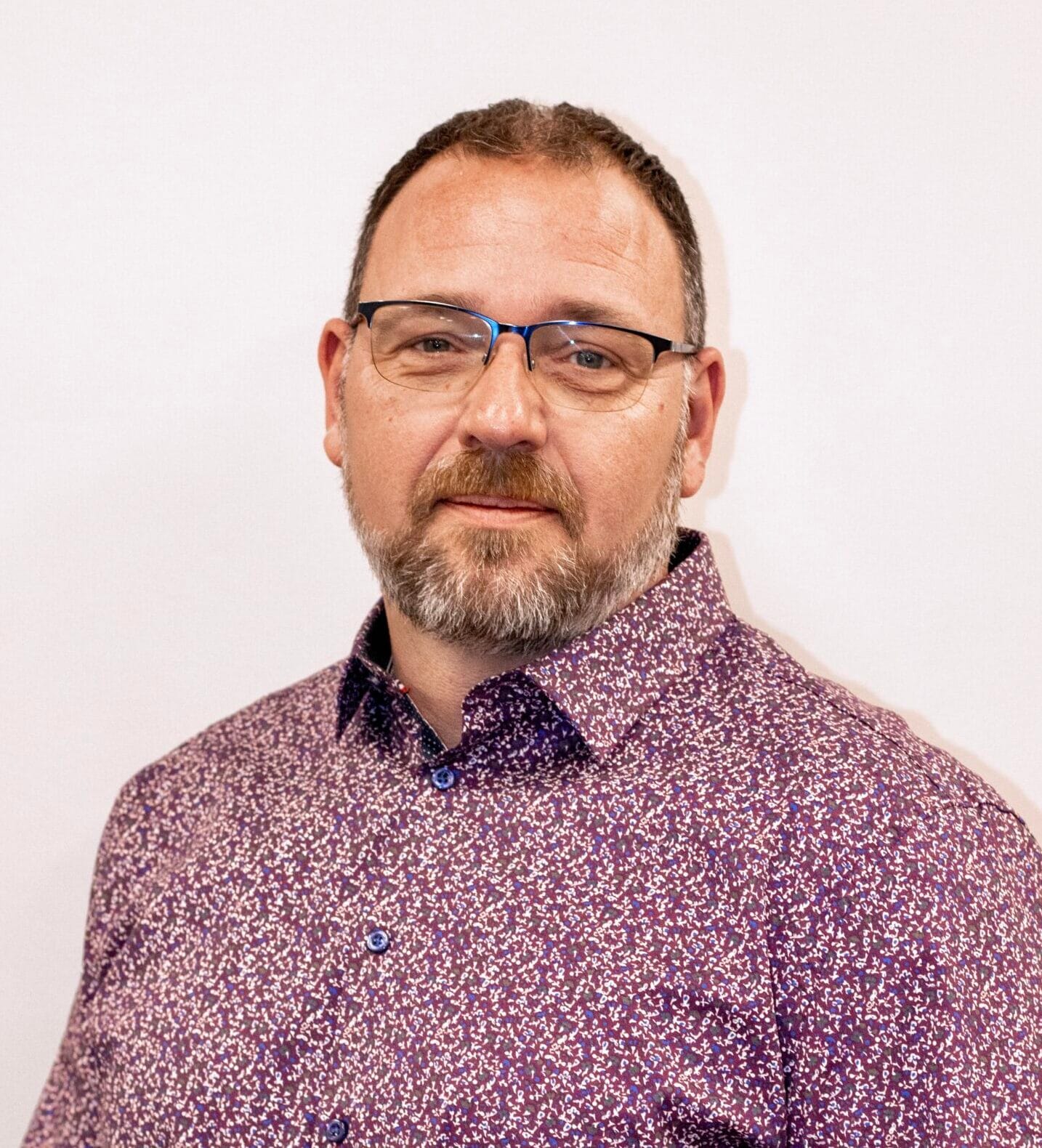 A man with a beard and eyeglasses wears a dark patterned shirt, pictured from the chest up against a plain white background.