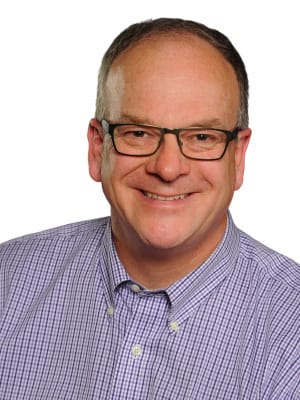 A man wearing glasses and a purple checkered shirt smiles widely against a plain white background.