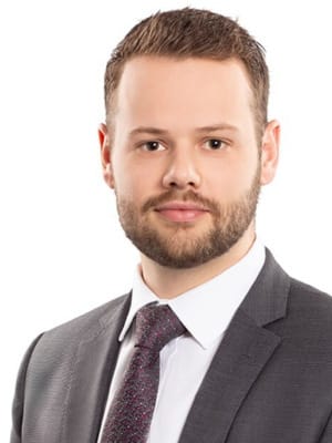 A man with a beard and short hair wears a suit and tie while looking directly forward against a white background.