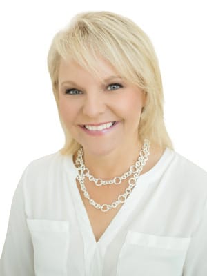 A smiling blonde woman with blue eyes wears a white collared shirt and a two strand silver chain necklace, captured against a white background.
