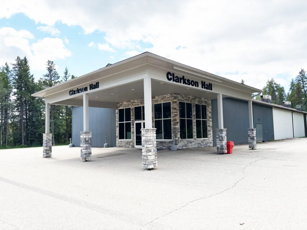 The Clarkson Hall building, featuring a stone and glass entrance with a covered portico, stands on a paved lot surrounded by trees under a partly cloudy sky.