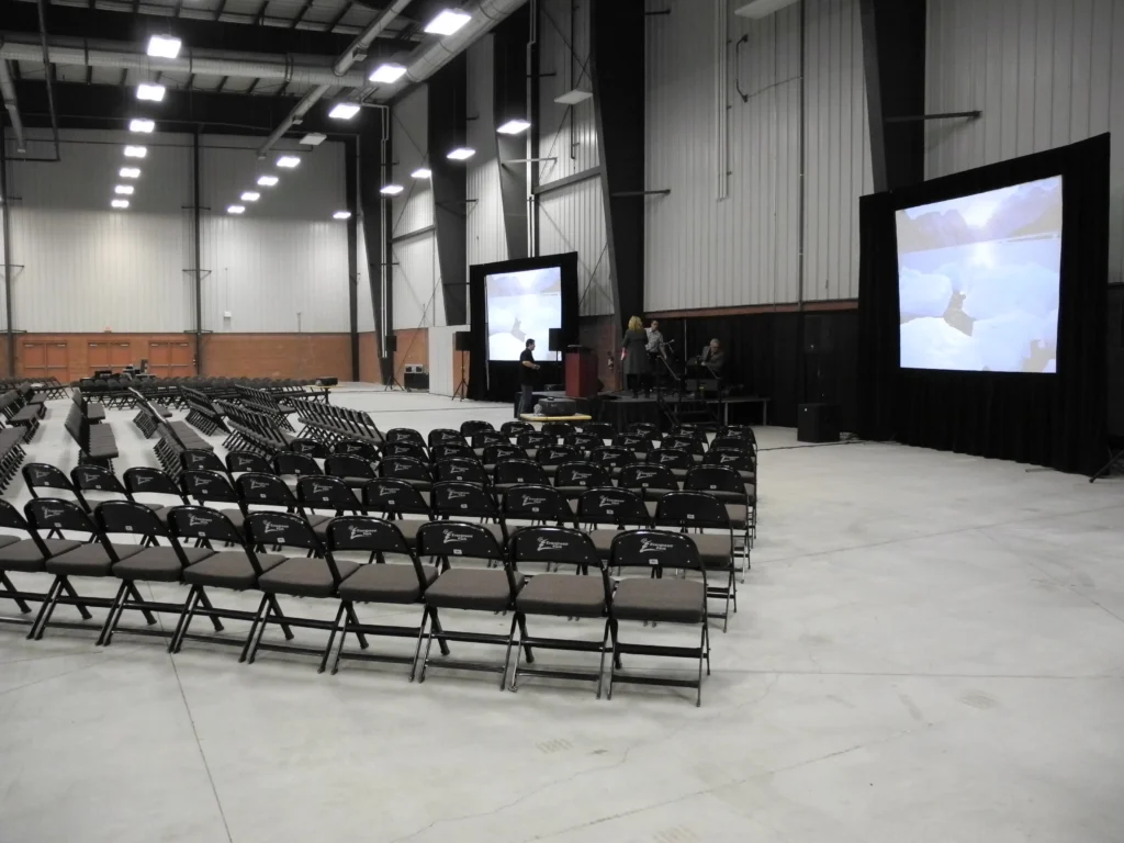 A spacious hall with rows of unoccupied dark folding chairs faces a stage where two large screens display a bright, snowy landscape, and a few people are visible around the setup.