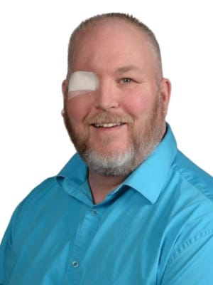 A smiling bearded man with a patch over his left eye wears a blue shirt against a white background.