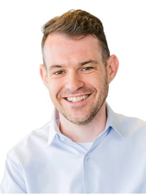 A smiling man with short hair and a light beard, wearing a light blue collared shirt, is shown from the chest up against a white background.