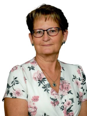 A woman with short dark hair and glasses wears a white top featuring a pink rose pattern and a pendant necklace, against a plain white background.