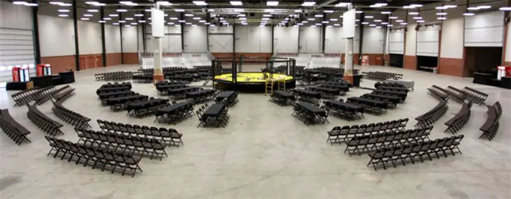 A large, empty arena is set up with a central circular fighting cage, numerous black tables and chairs arranged in curved sections for an audience, and concession stands along the perimeter.