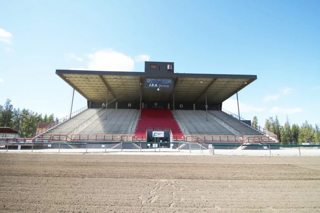 A covered grandstand with tiered concrete seating, a prominent red central section, and an upper observation booth displaying J.D.A. Raceway stands over a wide dirt track beneath a bright blue sky, bordered by trees.