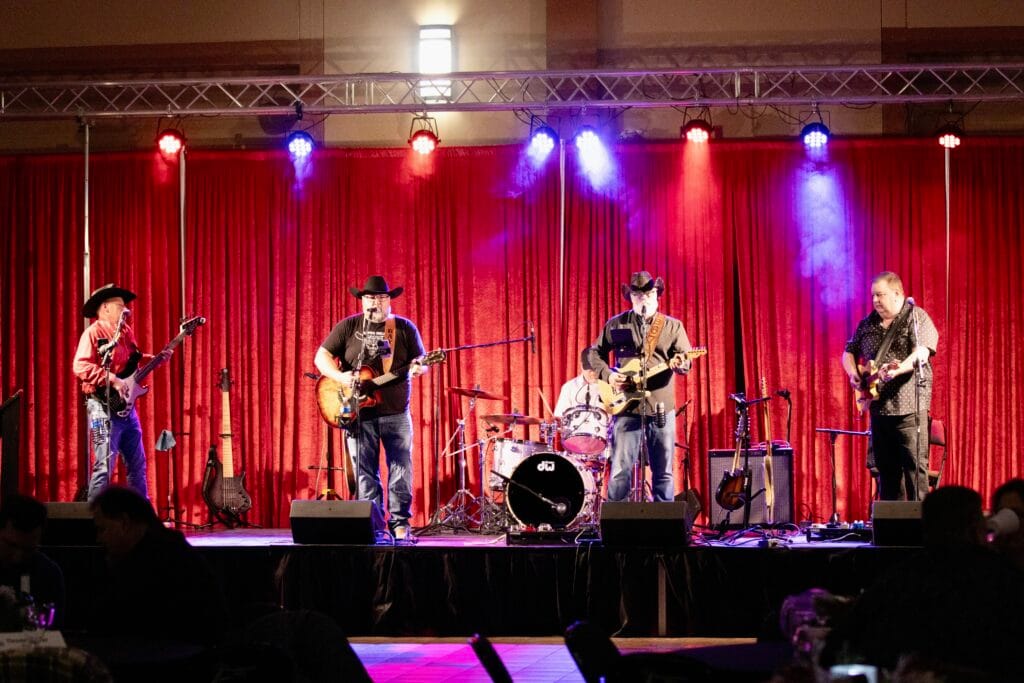 A band of five musicians, many wearing cowboy hats, performs on a stage lit by colorful red and blue lights, against a backdrop of red curtains, with an audience visible in the dark foreground.