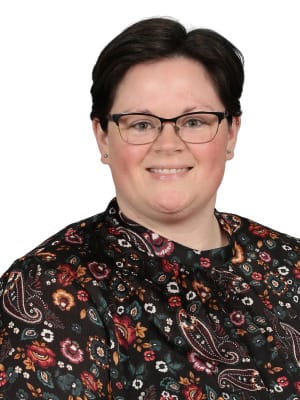 A smiling woman with short dark hair and glasses wears a dark patterned shirt against a plain white background.