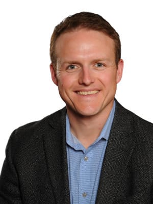 A smiling man with short brown hair wears a blue collared shirt and a dark blazer against a plain white background.