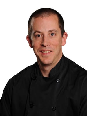 A man wearing a black chefs coat smiles slightly against a plain white background.