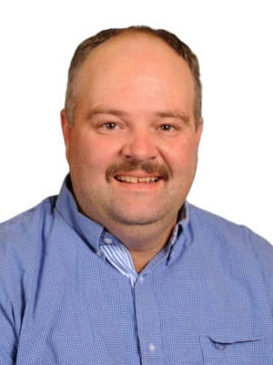 A man with a mustache, receding hairline, and a blue collared shirt smiles against a plain white background.