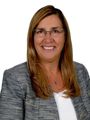 A smiling woman with long brown hair and glasses wears a gray and white textured jacket over a white top against a plain white background.
