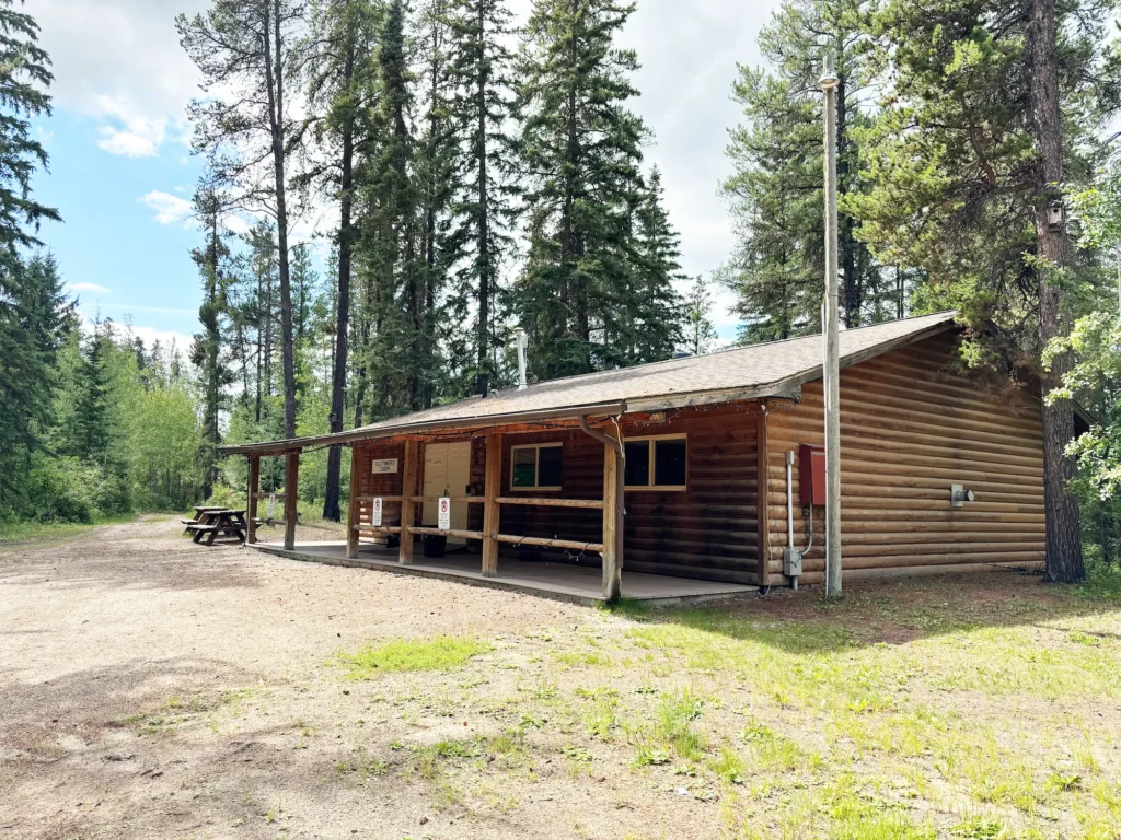 A log cabin with a covered porch and a picnic table stands on a dirt clearing, surrounded by tall evergreen trees under a partly cloudy sky, with a utility pole nearby.