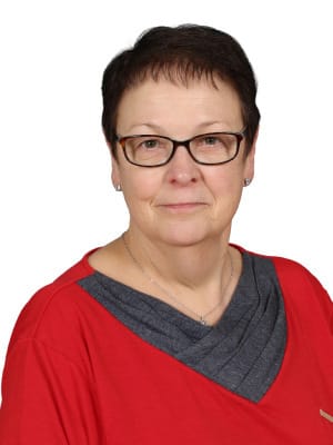A smiling woman with short dark hair and glasses wears a red shirt with a gray V neck detail against a white background.