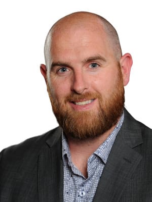 A smiling man with a shaven head and a reddish brown beard wears a patterned button down shirt and a dark blazer against a plain white background.
