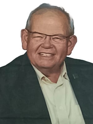 An older man wearing glasses, a light shirt, and a dark jacket smiles at the camera against a white background.