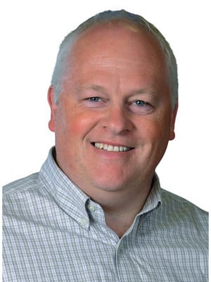 A man with short gray hair and a plaid shirt smiles, revealing his teeth, while looking towards the viewer on a white background.