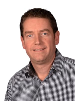 A man with short brown hair and a slight smile wears a black and white checkered shirt against a plain white background.
