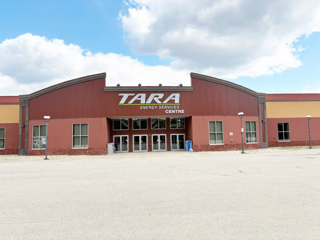 A large reddish brown building featuring the sign TARA ENERGY SERVICES CENTRE stands with numerous windows and glass doors, under a blue sky dotted with white clouds, with a spacious paved area in front.