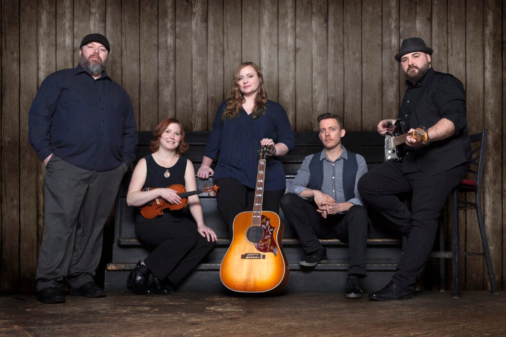 Five musicians, three men and two women, pose with an acoustic guitar, a violin, and an electric guitar in a room with a rustic wooden wall and floor.