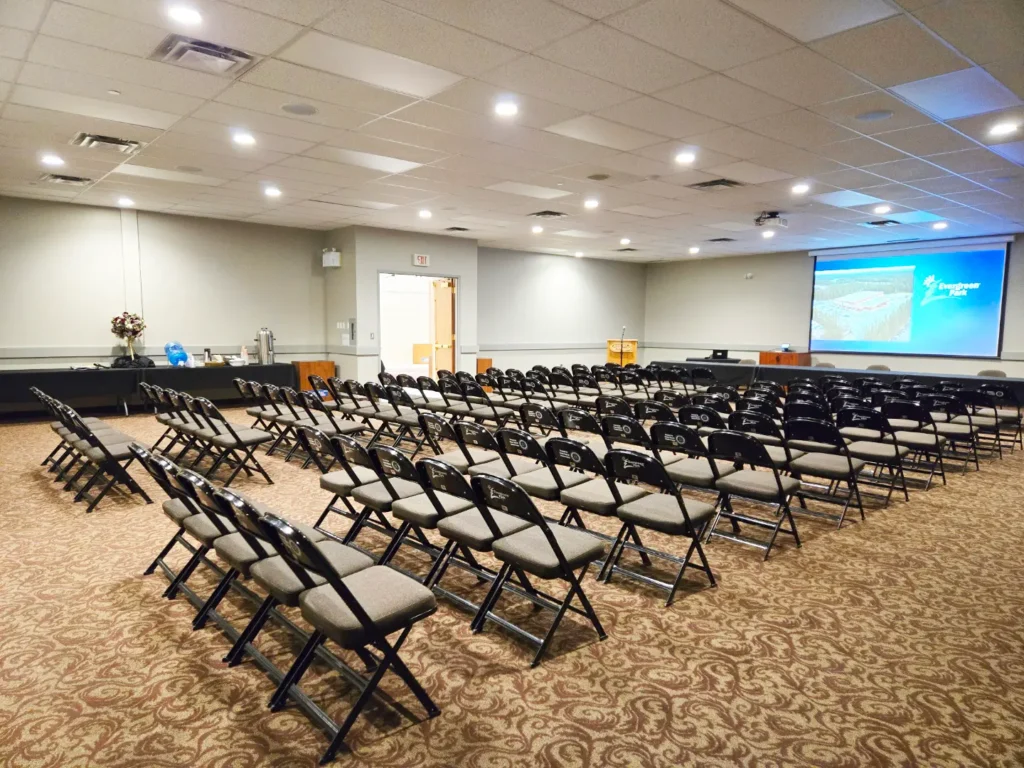 Numerous empty folding chairs are neatly arranged in rows on a patterned carpet within a conference room, facing a large projection screen displaying an image and the words Evergreen Park, with a refreshment station and an exit door visible along the walls.