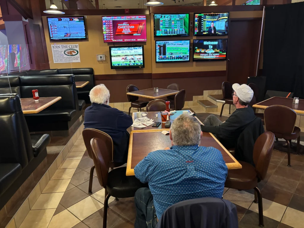 Three men are seated with their backs to the viewer at tables in a casual dining area featuring multiple television screens displaying horse racing and casino programs.