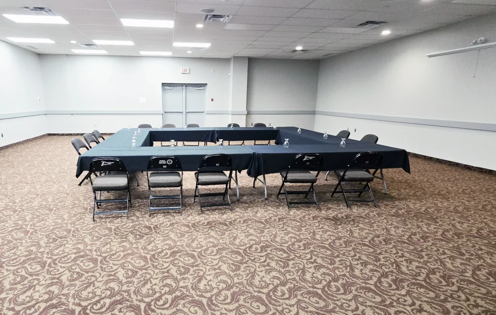 A brightly lit conference room features a large U shaped table covered in black tablecloths with chairs and water glasses set upon a patterned carpet, with plain walls and double exit doors in the background.