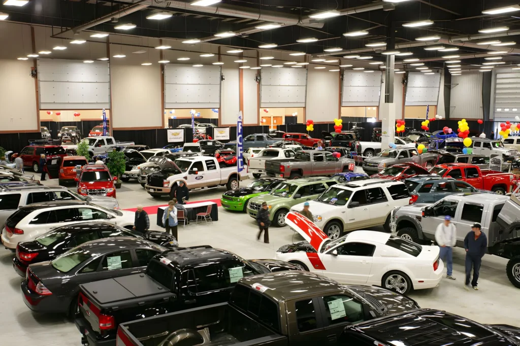 Numerous cars, trucks, and SUVs are displayed in a large, brightly lit indoor venue, with people walking around and observing the vehicles, some of which have their hoods open.