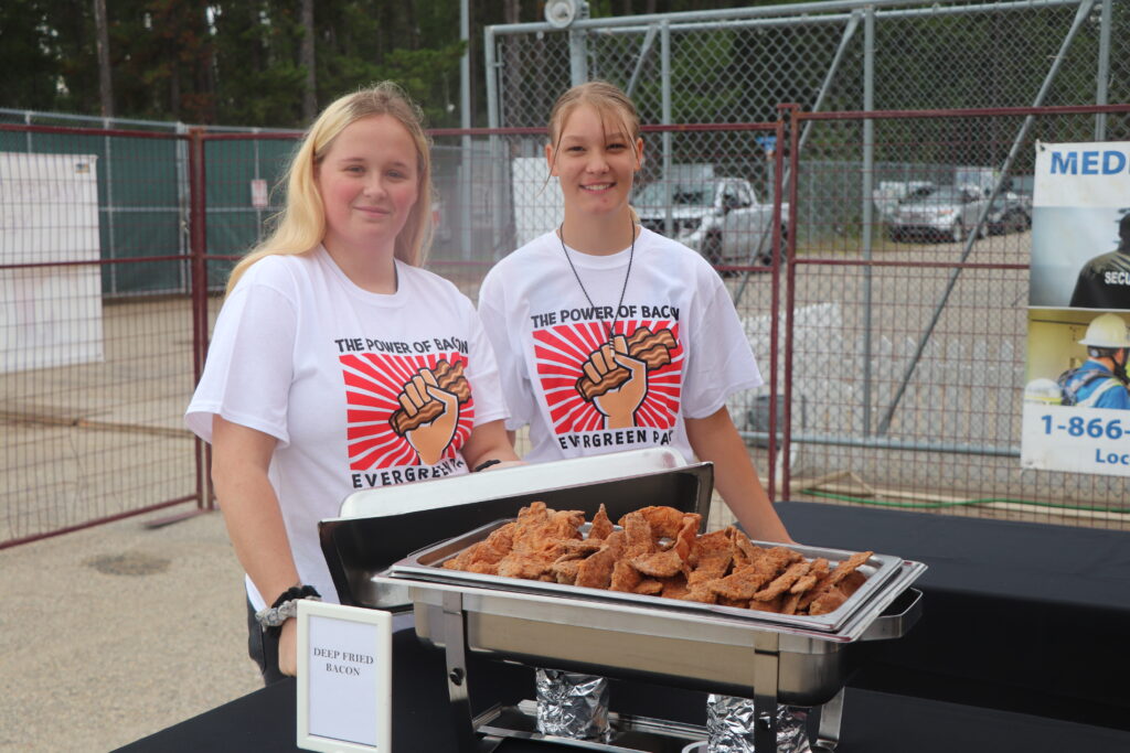 Two young women, wearing matching white shirts emblazoned with The Power of Bacon, stand behind a table featuring a chafing dish filled with deep fried bacon and a sign explicitly labeled Deep Fried Bacon, all set outdoors against a backdrop of a fence and trees.