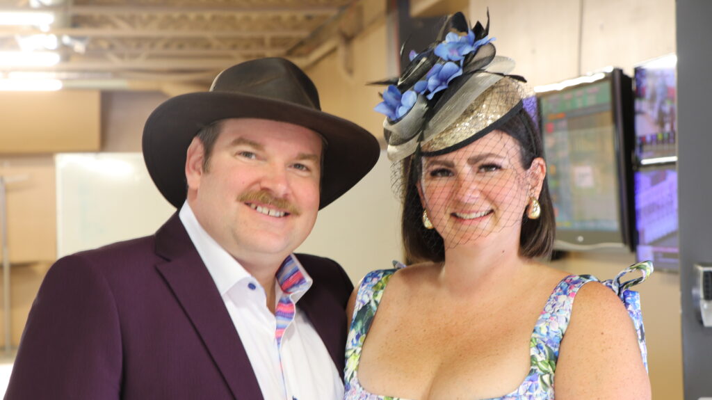 A smiling man wearing a dark fedora and purple suit jacket stands beside a woman in a colorful floral dress and an ornate fascinator with a face veil and blue flowers.
