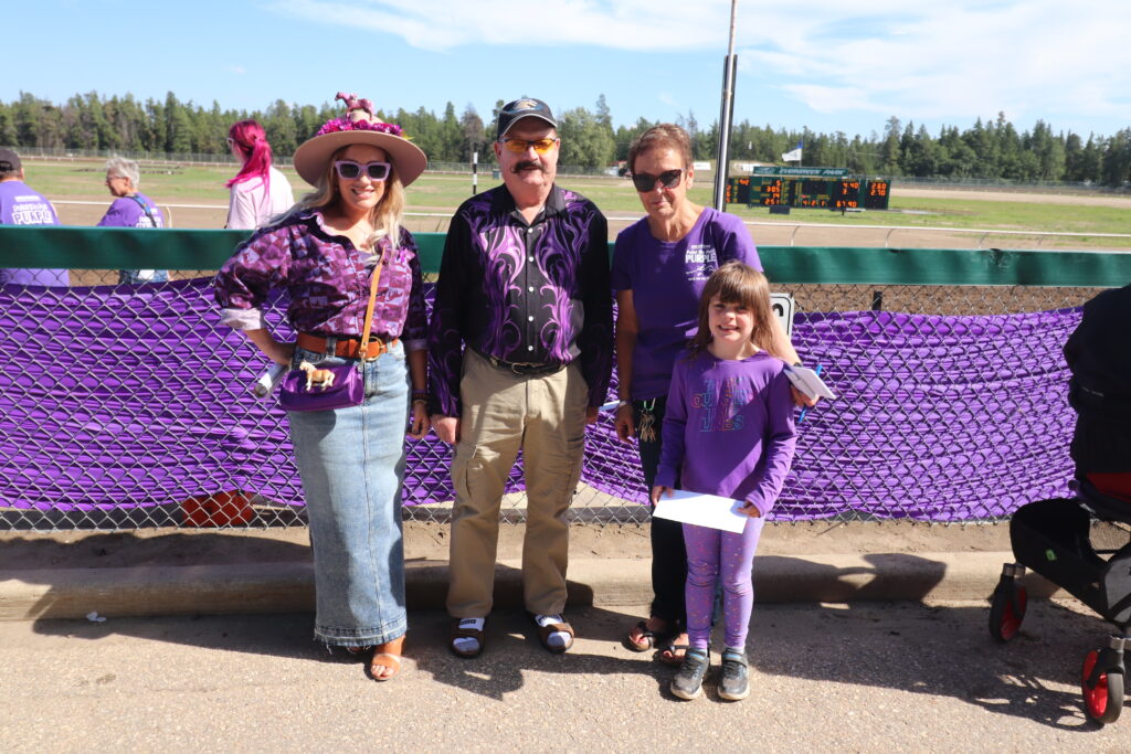 A smiling group of four people, predominantly dressed in purple and including a man with a mustache and a girl holding a paper, poses in front of a purple draped chain link fence at a sunny outdoor racetrack.