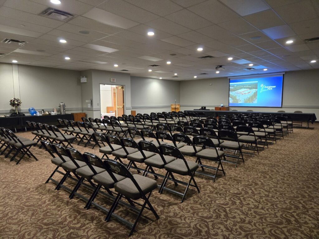 This wide shot captures an empty conference room with numerous rows of black and gray folding chairs facing a large projection screen that displays an image and the words Evergreen Park, alongside a refreshment table to the left and an exit door visible on the right.