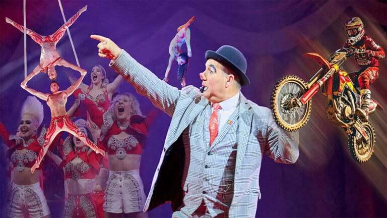 A clown in a plaid suit points excitedly amidst a lively circus scene featuring multiple acrobats, a woman suspended by her hair, a motocross rider, and cheering performers on a purple stage.