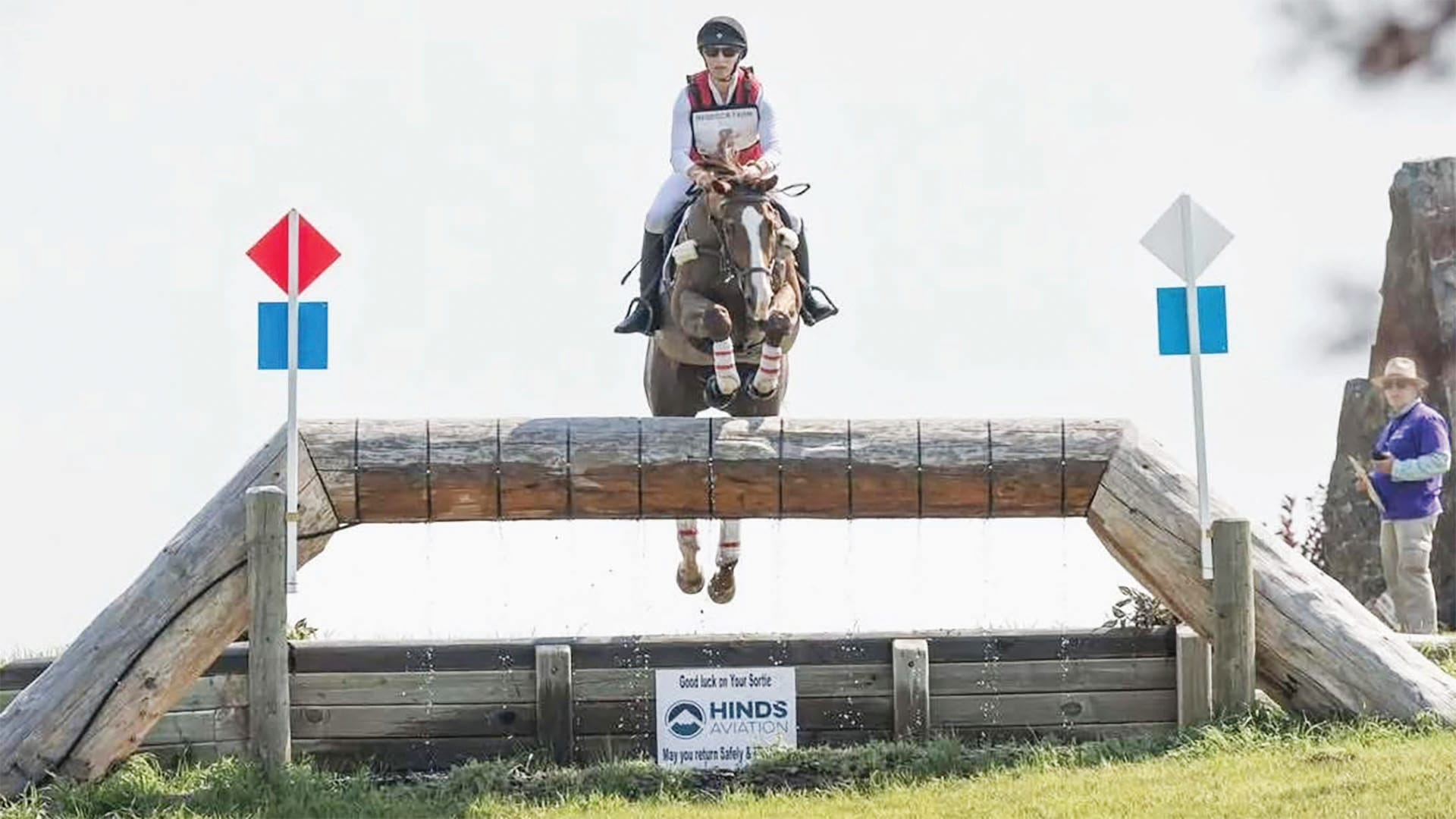 A rider atop a brown horse is captured midair as they jump over a robust wooden log fence, flanked by red and blue marker flags, while an observer watches nearby.