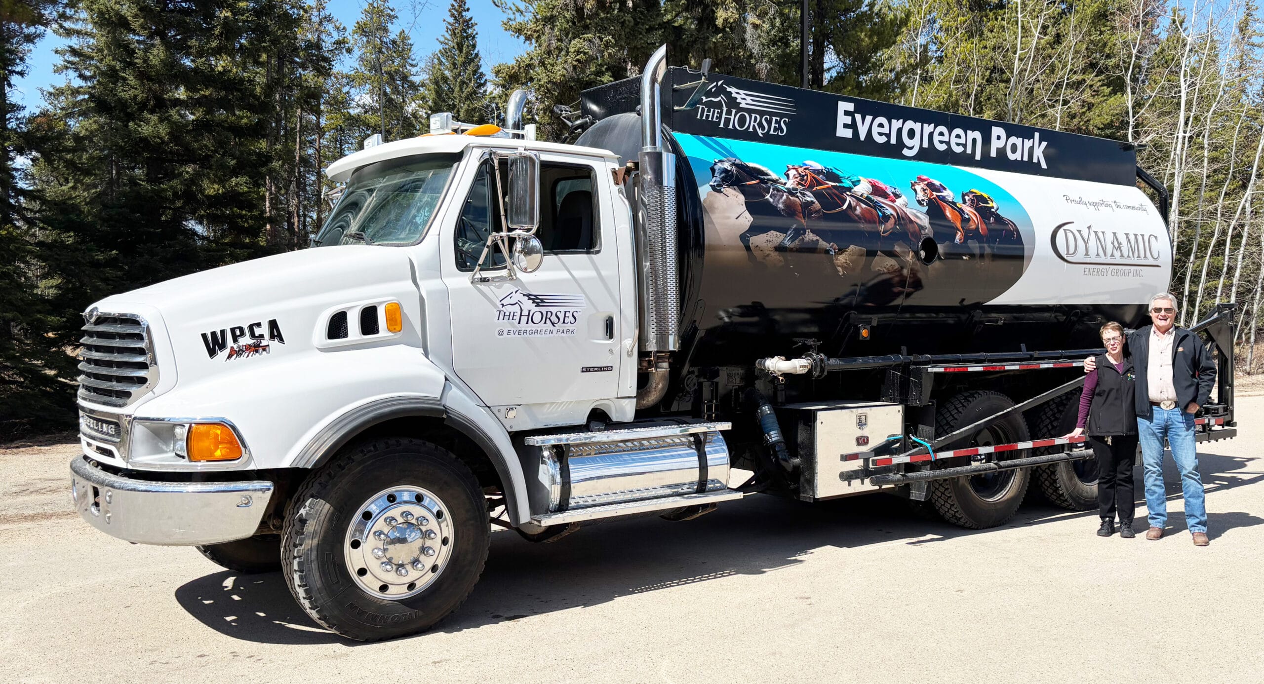 A white tanker truck with a black tank displaying large graphics of racehorses and the name Evergreen Park is parked on a paved lot, with a smiling man and woman standing next to it under a clear blue sky and green trees.